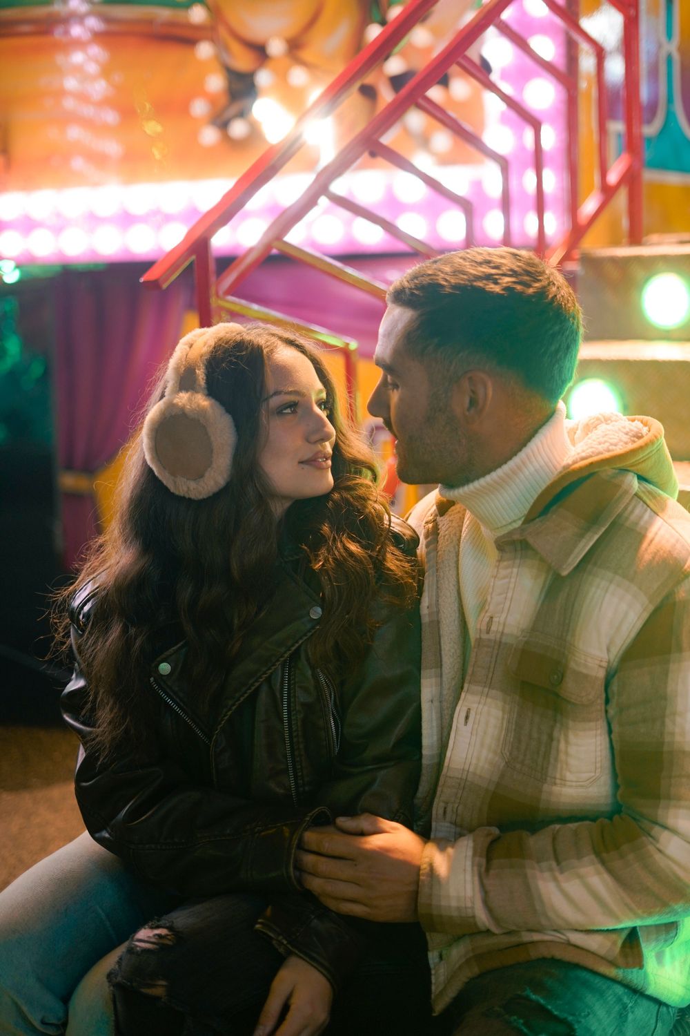 Romantic couple at carnival with woman wearing earmuffs amid colorful neon lights.