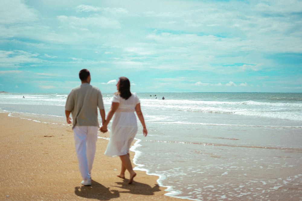 A couple in white clothing walks hand in hand along a sandy beach at the ocean's edge.
