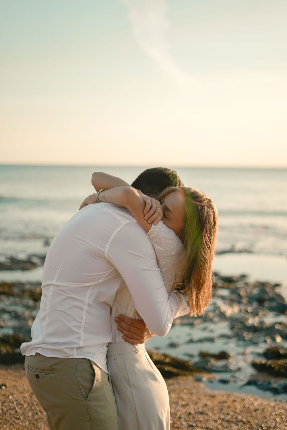 Romantic couple embracing on a beach at sunset wearing white clothes against an ocean backdrop.