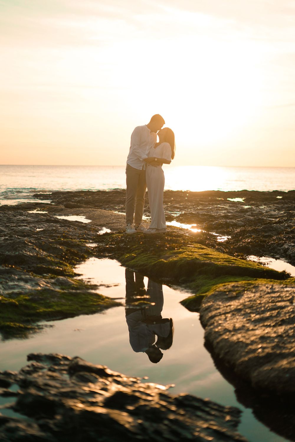 A silhouetted couple embracing on rocky coastline during golden sunset with reflection in tide pool.