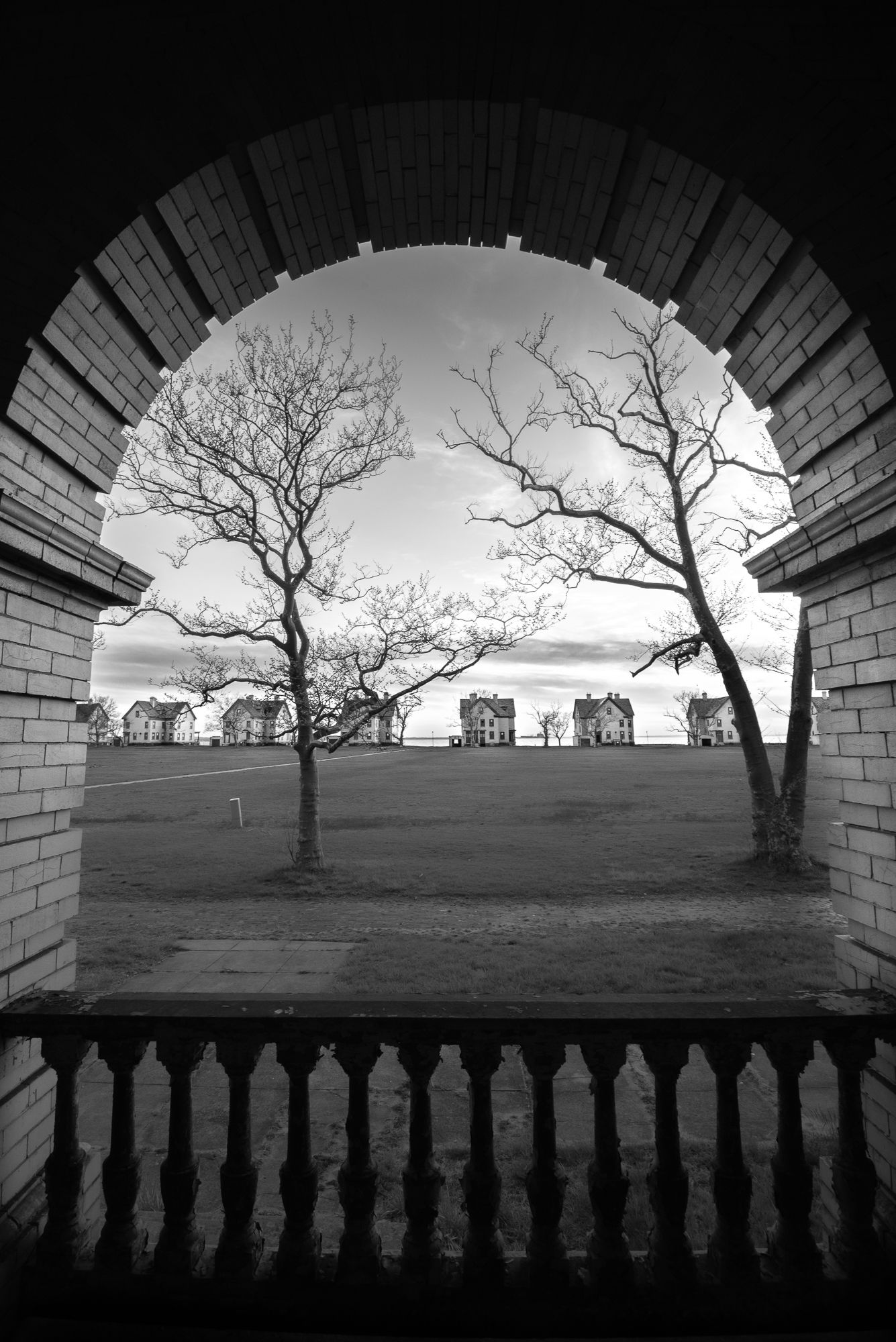 Black and white photograph of Officers Row through stone archway of bare trees and buildings on horizon Fort Hancock, Sandy Hook, NJ