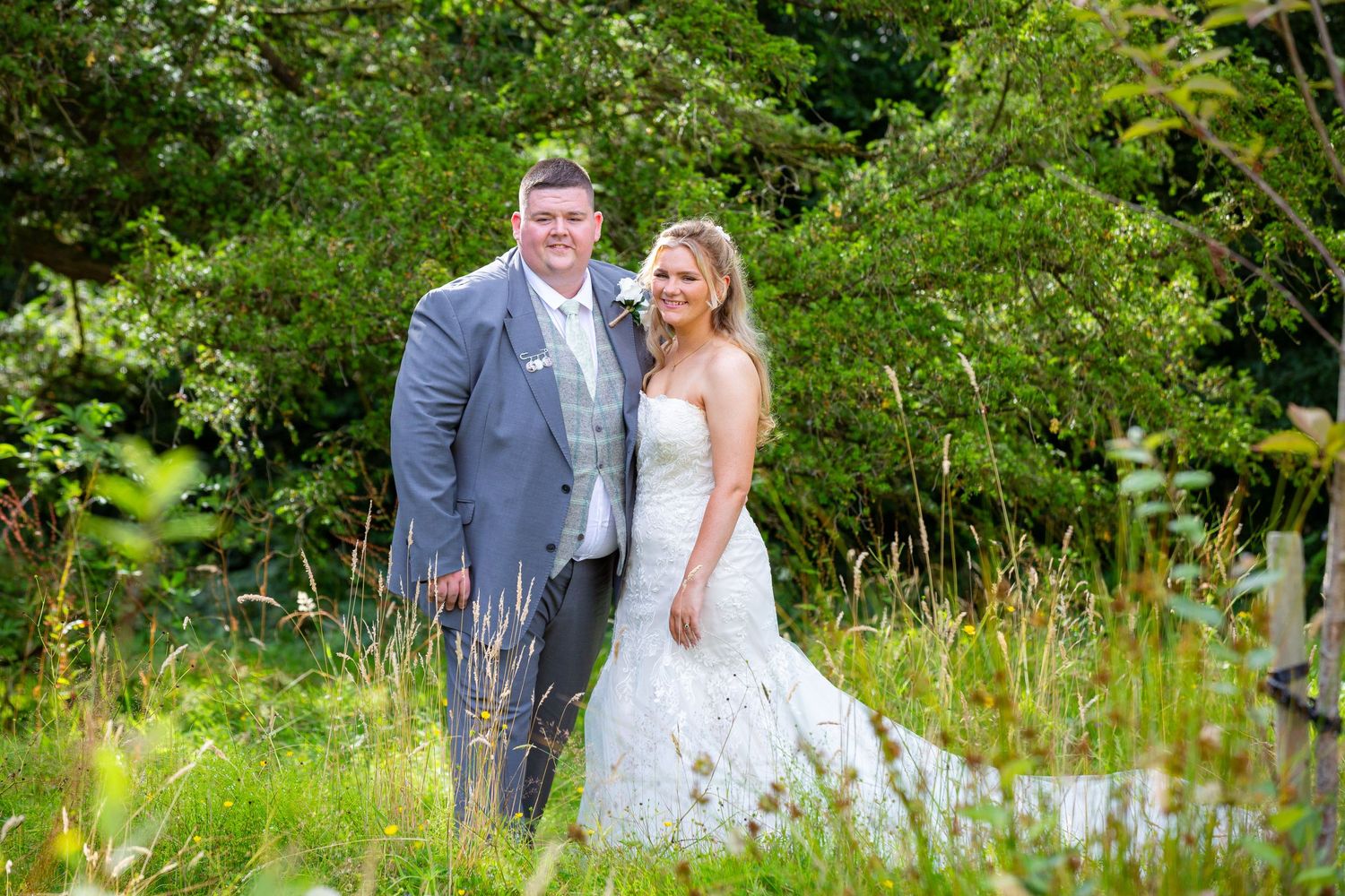 Bride and groom smile together in a lush green outdoor setting on their wedding day.