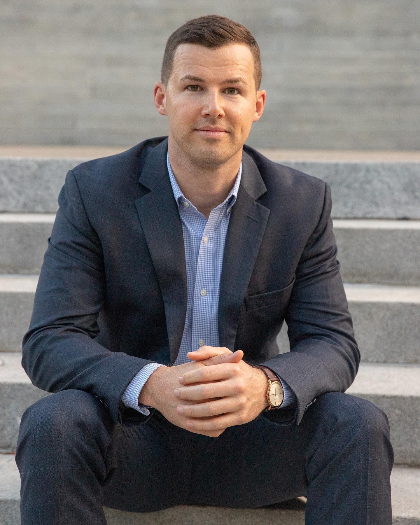 Professional sitting on outdoor stone steps wearing navy suit and light blue dress shirt with hands clasped.