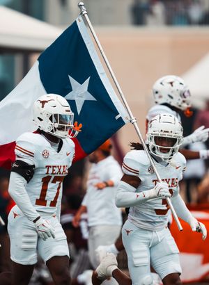 Texas Longhorns players in white uniforms run onto the field carrying Texas state flags.