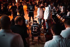 Basketball player wearing jersey number 12 kneels on court during pre-game introductions amid dramatic arena lighting.