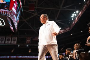 Coach in white outfit stands courtside during a basketball game in a packed indoor arena.