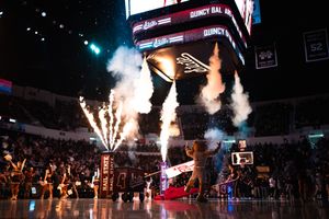 Indoor basketball arena pregame show with pyrotechnics, fireworks, and cheerleaders under a large scoreboard.