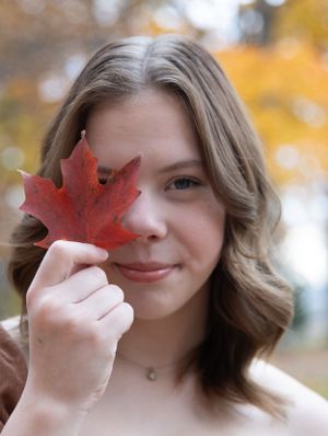 Someone holds up a red maple leaf against an autumn backdrop with bokeh effects.