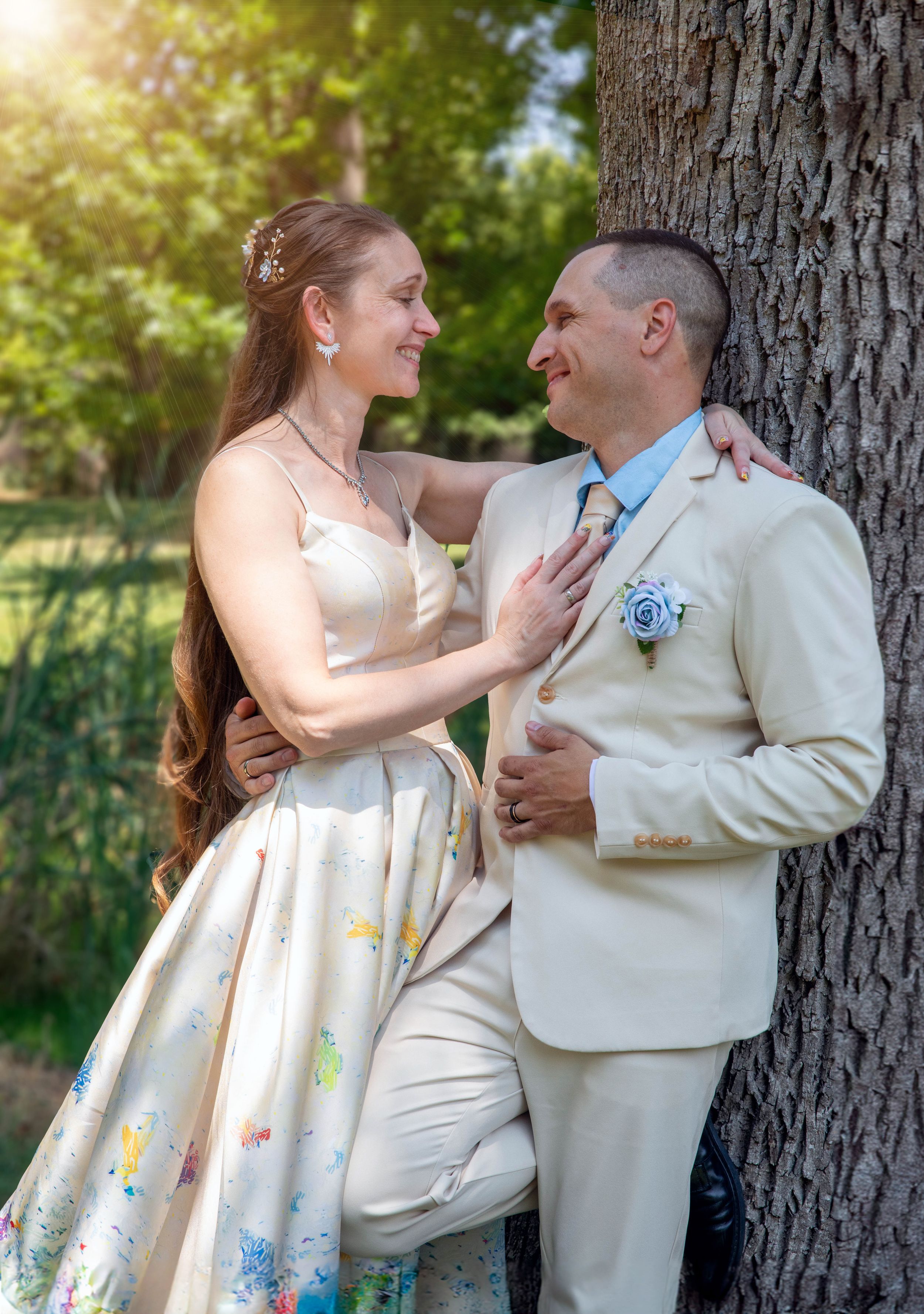 A couple in matching cream formal attire poses against a tree trunk in dappled sunlight.