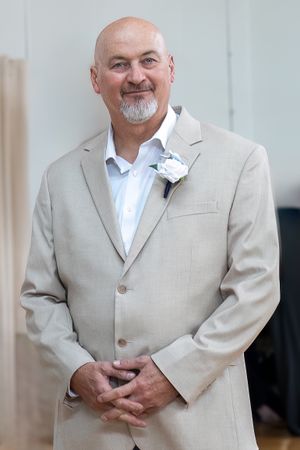 Gentleman in light beige suit with white boutonniere smiles during formal event.