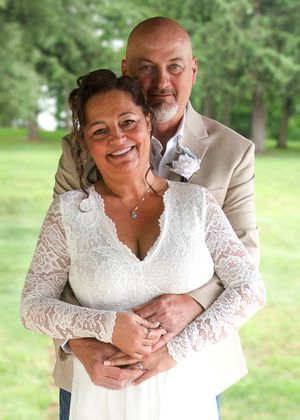 A happy couple embraces outdoors on their wedding day, with the bride wearing a white lace dress and groom in tan suit.