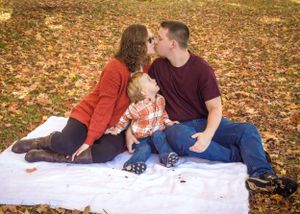 A family sits on a white blanket surrounded by fallen autumn leaves.