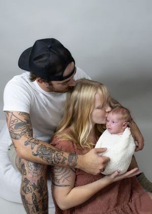 Intimate family portrait with tattooed parent cradling newborn wrapped in white.