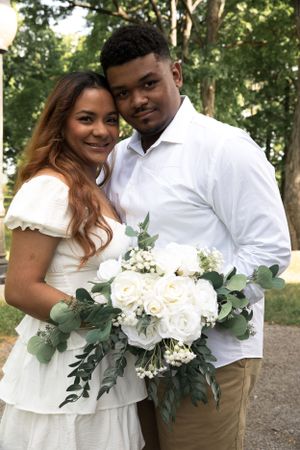 A couple poses with a beautiful white rose and eucalyptus wedding bouquet in an outdoor setting.