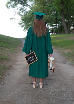 Graduate in emerald green cap and gown holds small chalkboard while standing on dirt path.