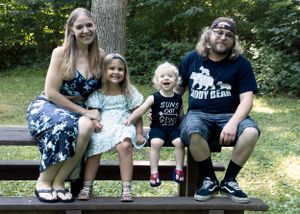 Family of four sitting together on wooden bench in wooded outdoor setting.