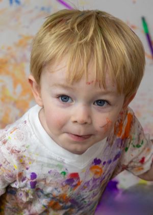 Paint-covered child smiling during messy art activity with colorful background.