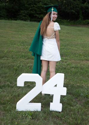 Graduate in green cap and gown poses on white numbers 24 during outdoor photo session.