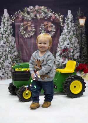 A holiday photo setup with toy John Deere tractor against a rustic winter backdrop with wreaths and snow.