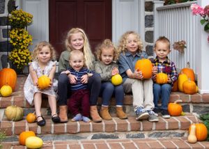 Children sitting on brick steps holding pumpkins with fall decorations and flowers in the background.