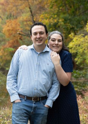 A couple embraces outdoors with autumn foliage in the background.