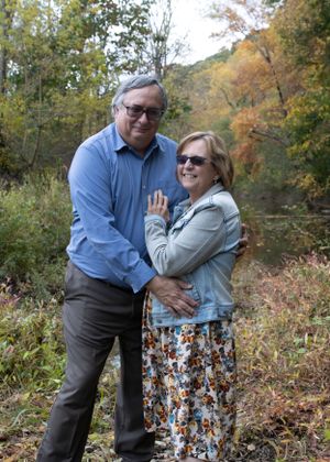 A couple poses together in a scenic fall setting with colorful trees behind them.