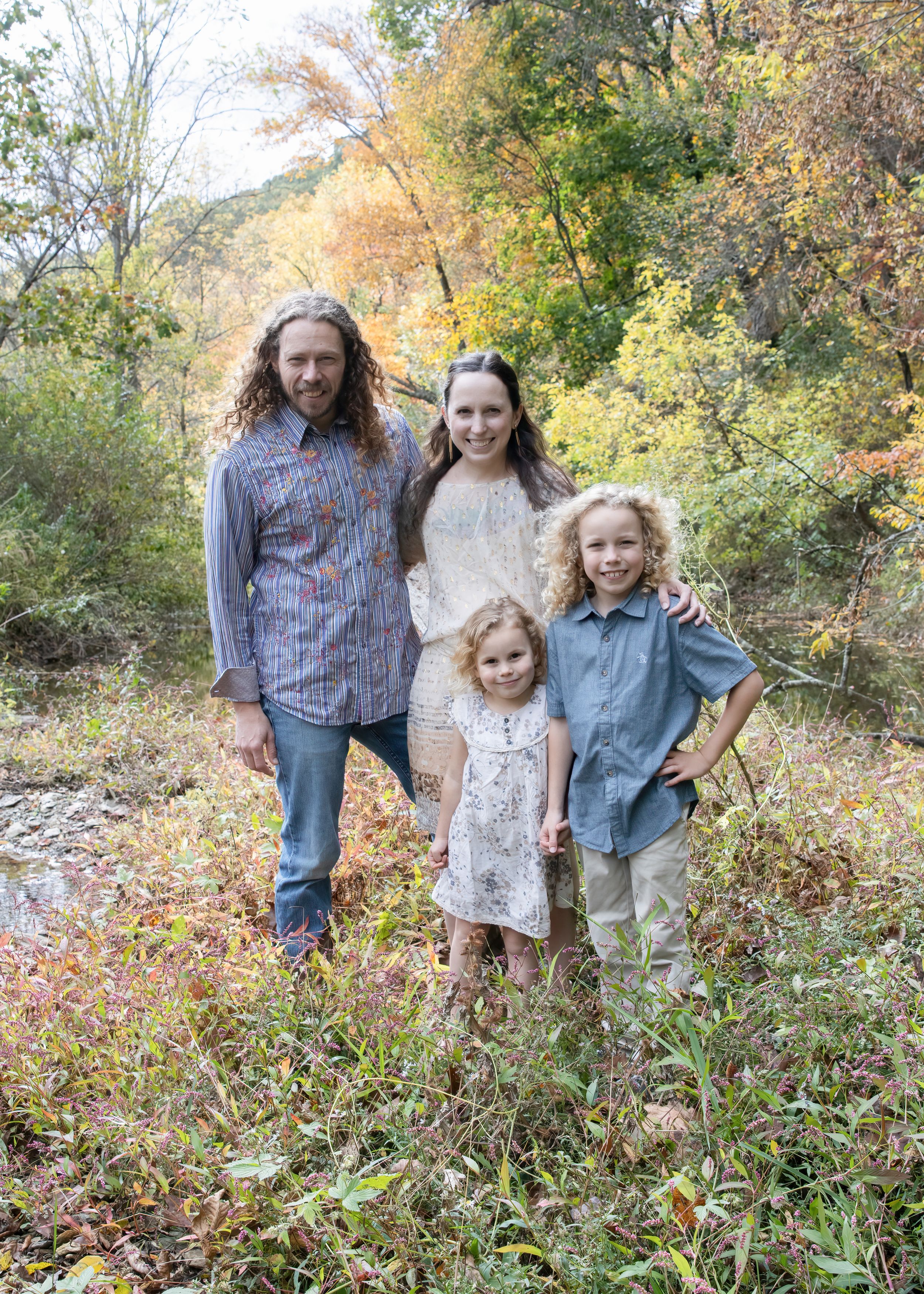 A family of four poses together outdoors during autumn with fall foliage in the background.