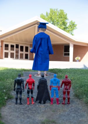Graduate in blue cap and gown standing with superhero action figures in front of school building.