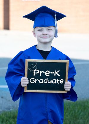 Young graduate in blue cap and gown holds chalkboard sign reading Pre-K Graduate.