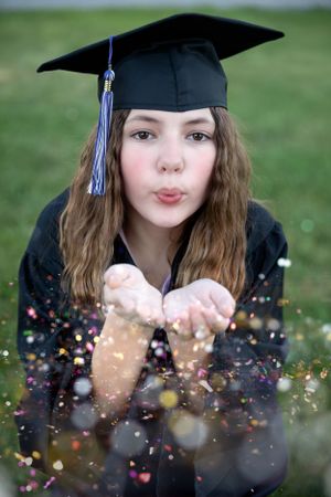 Graduate in black cap and gown blows glitter and confetti during outdoor photo shoot.