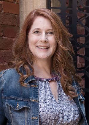 Portrait in casual denim jacket and patterned top against brick wall background.