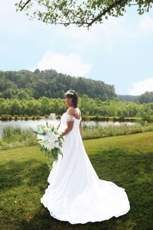 Bride in elegant white wedding dress with long train holding bouquet poses near lake and rolling hills.