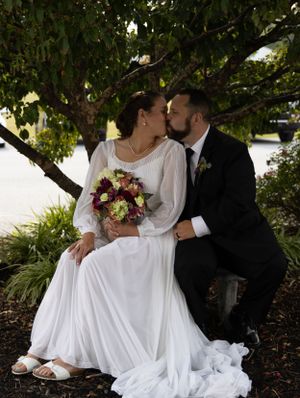 Romantic wedding couple sitting on bench under tree branches near waterfront.