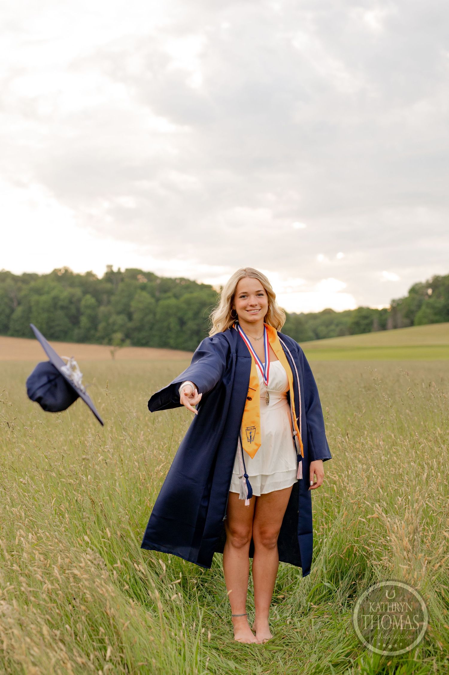 Graduate stands joyfully in a field wearing a navy graduation gown and academic regalia during golden hour.