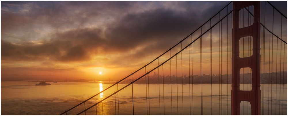 Golden sunset silhouettes the iconic Golden Gate Bridge cables against a dramatic orange sky over San Francisco Bay.
