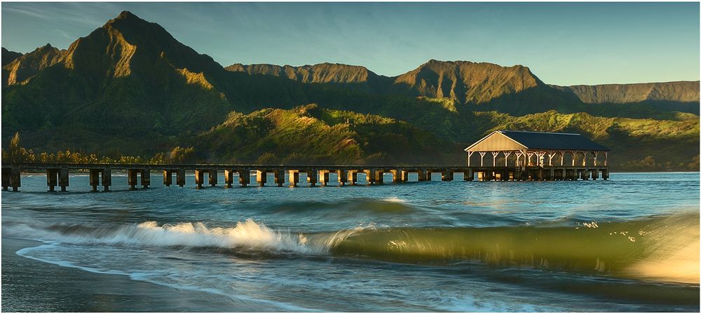 Waves crash near Hanalei Pier on Kauai's north shore with rugged Na Pali mountains rising in the background.
