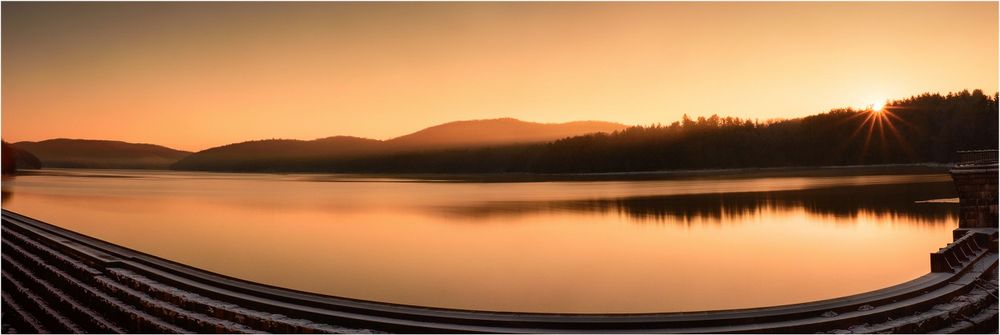 Panoramic view of a serene lake at sunset with orange sky reflecting in calm waters between silhouetted mountains.
