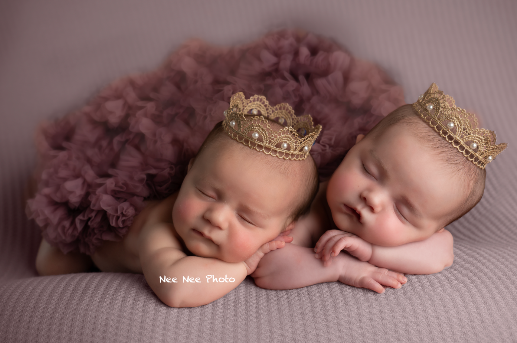 Two sleeping newborn twins wearing gold lace crowns rest on a purple tulle skirt in a professional photo. Embracing Life’s Beauty