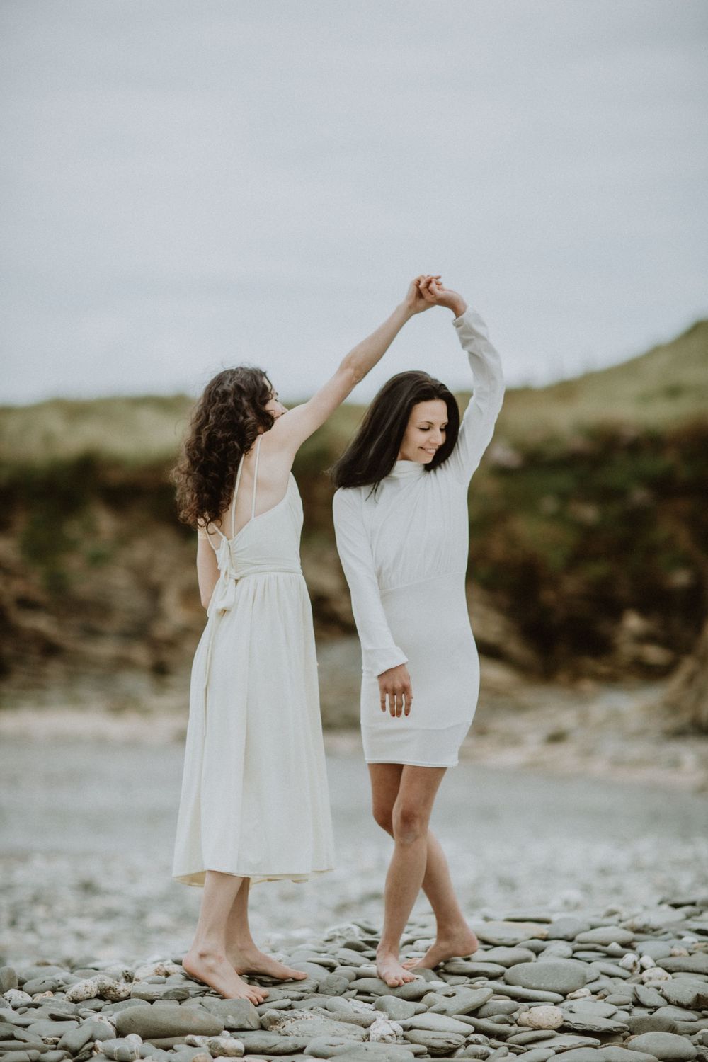 Two people in white dresses dancing on rocky beach with cliffs in background.
