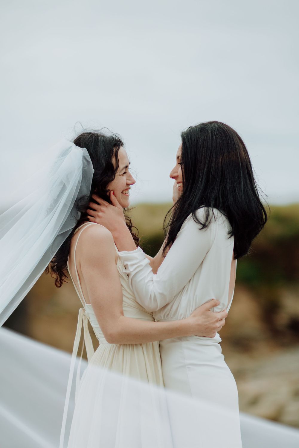 Wedding moment on rocky beach with veil flowing in coastal breeze.