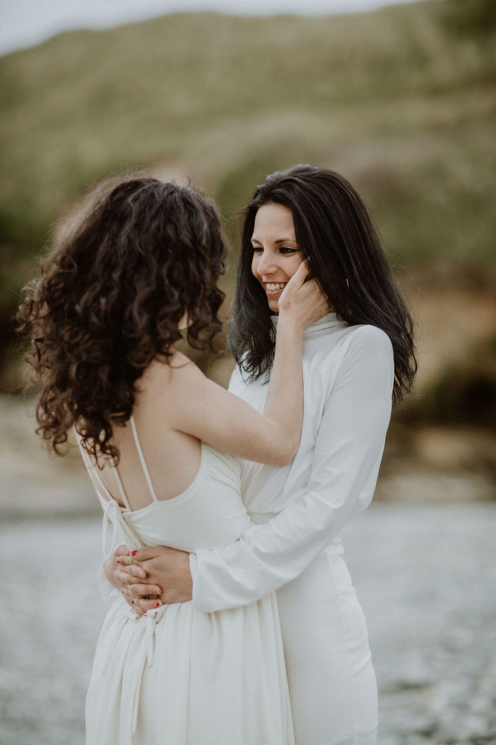 Two people in white dresses share an intimate moment on a rocky beach at sunset.