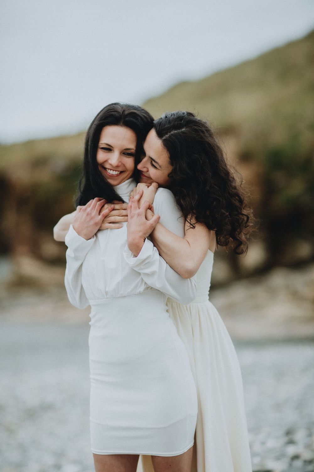 Two people in white dresses share a joyful embrace on a rocky coastline.