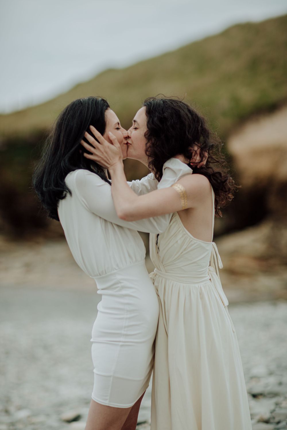 Romantic moment between two people in white dresses kissing on a rocky beach.