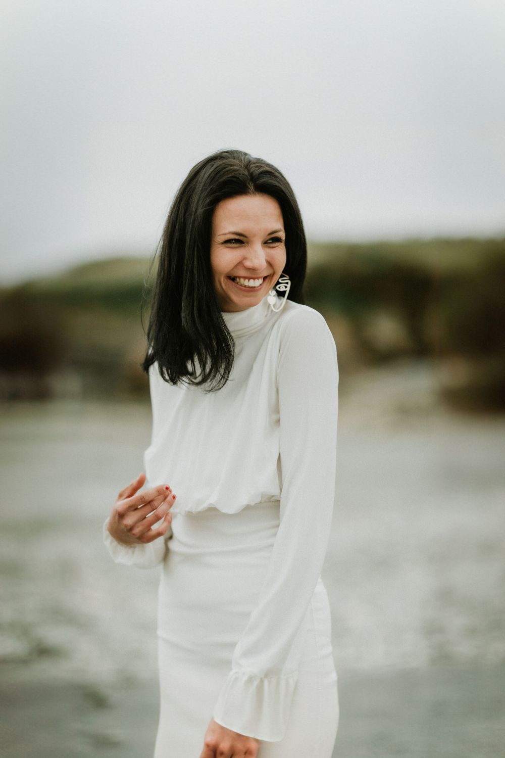 Person in a long-sleeved white dress smiling brightly on a cloudy beach.