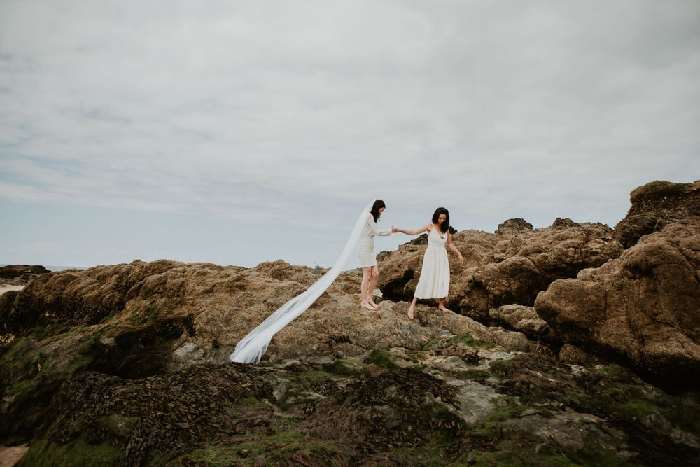 Two people in white dresses stand atop rocky cliffs with flowing fabric in the wind.