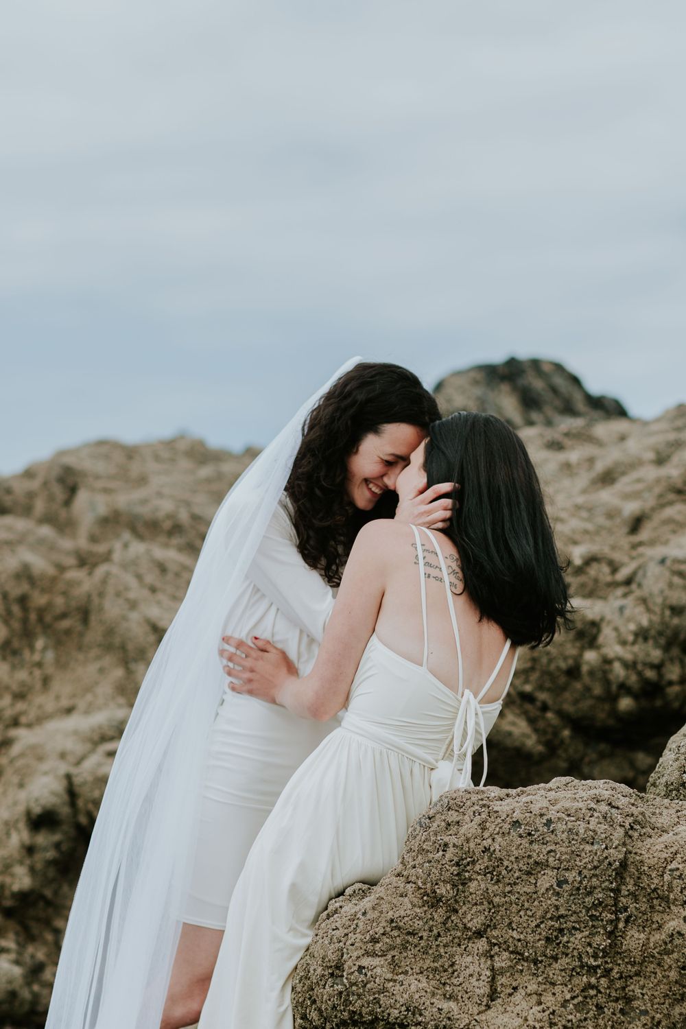 Couple in white dresses sharing an intimate moment among coastal rocks.