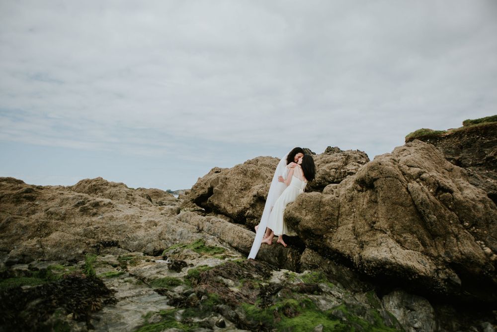 Person in white dress standing on mossy rocks against cloudy coastal backdrop.