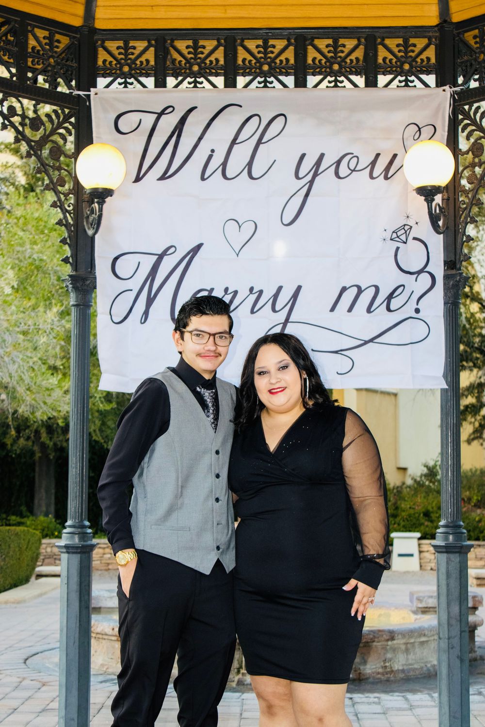 Newly engaged couple smiling together under Will You Marry Me banner in ornate gazebo.