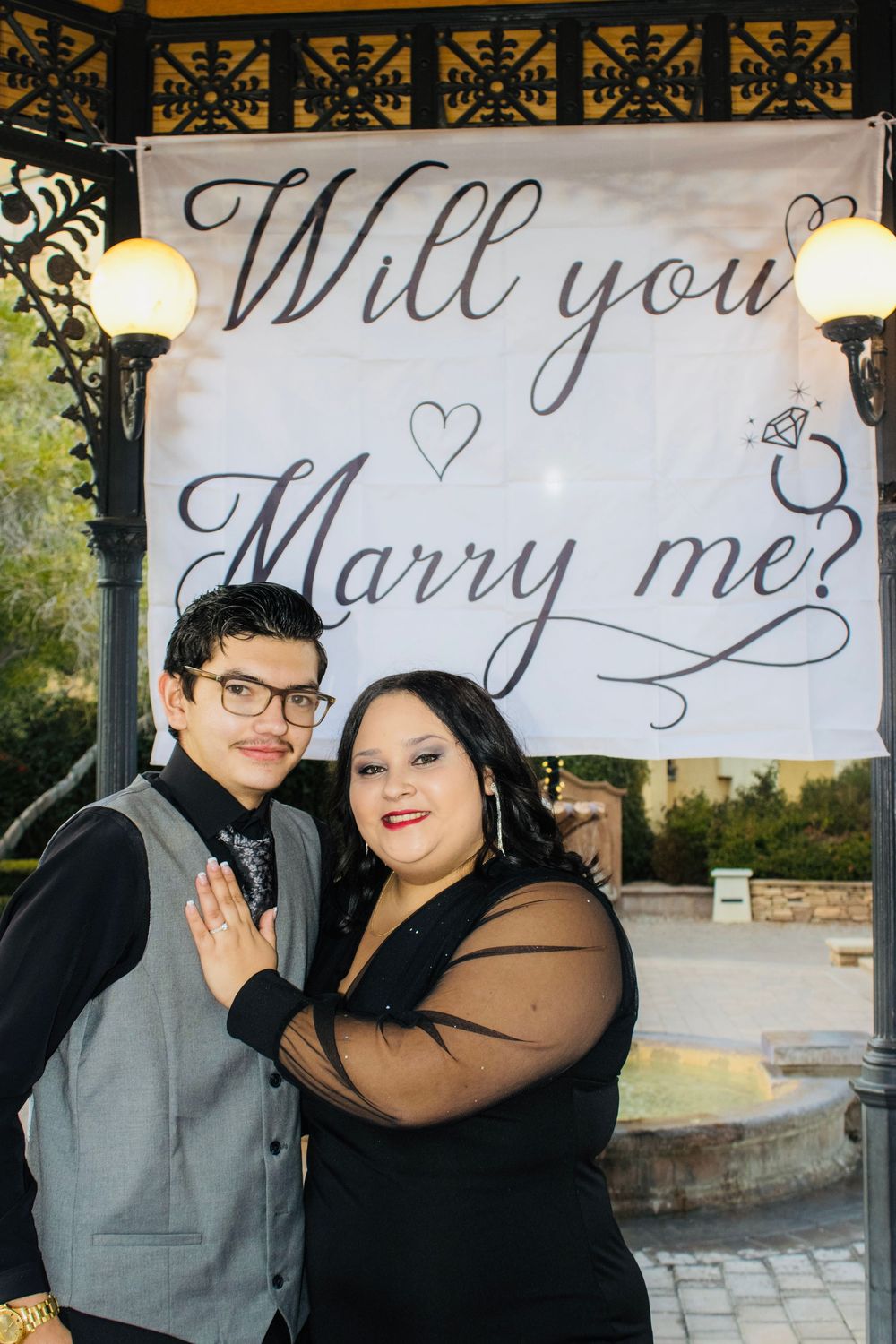 Happy couple posing together under Will You Marry Me banner in decorated gazebo setting.