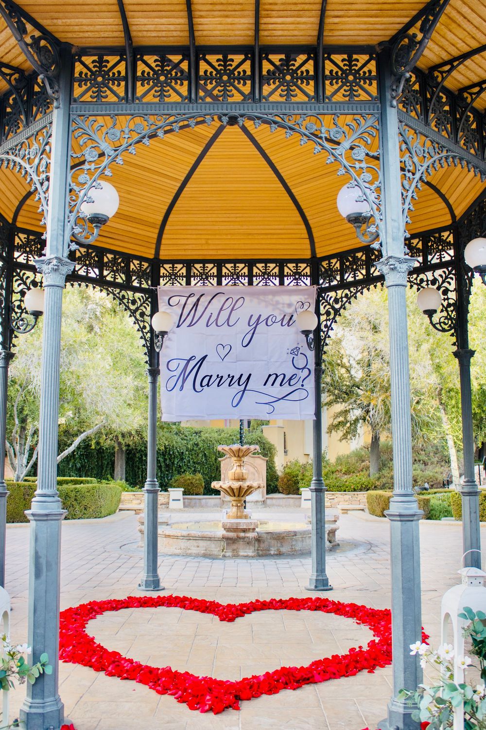 Ornate gazebo decorated with a red rose petal heart and a Will You Marry Me banner inside.